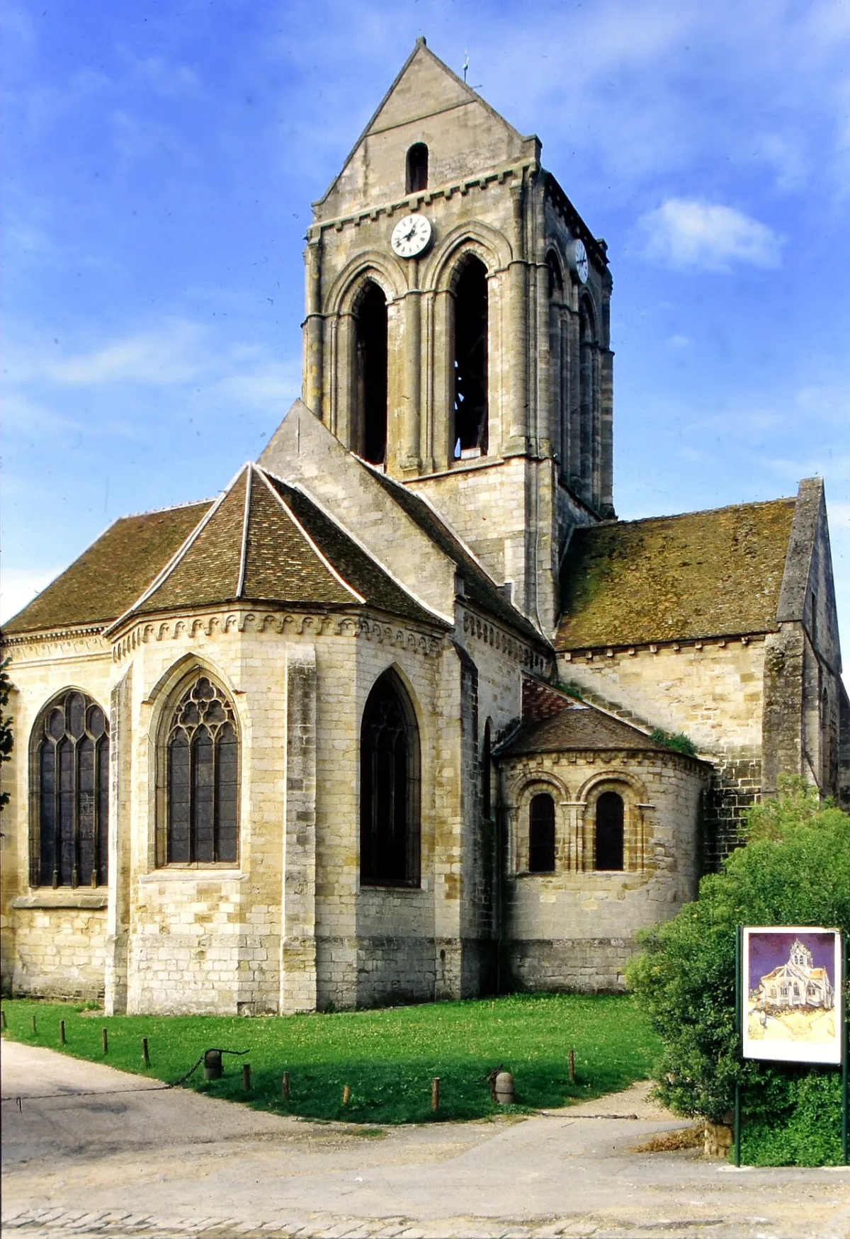 De kerk van Auvers-sur-Oise gezien vanaf het koor, met naast het gazon een bordje met een reproductie van Van Goghs schilderij van dezelfde kerk