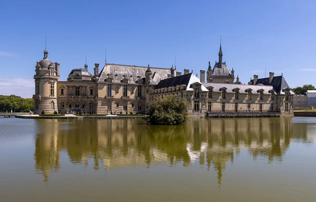 Zijaanzicht van het Château de Chantilly met de weerspiegeling van het kasteel in het omringende water, onder een blauwe lucht